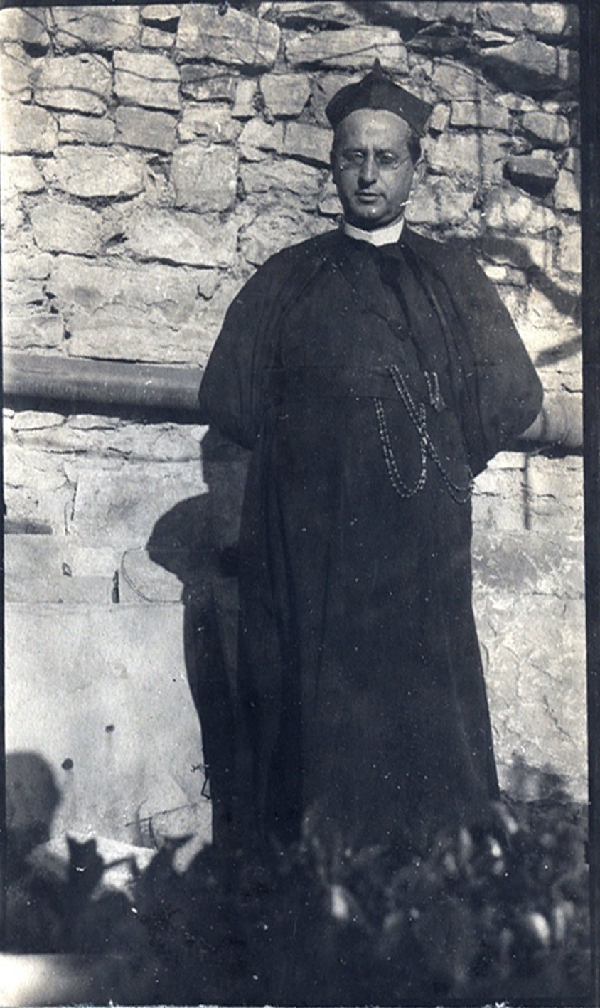 Full-length photograph of Fr Donato Jiménez in cloak and biretta, in front of a stone wall.
